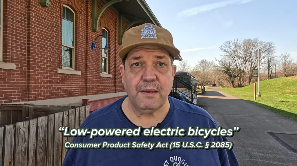Front-view of a man in a brown cap outside a brick building; on-screen text reads 'Low-powered electric bicycles' and 'Consumer Product Safety Act' at the bottom.